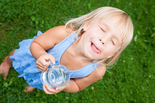 High angle view shot of blonde little girl wearing blue dress holding glass of water licking her lips with her eyes closed in a summer garden