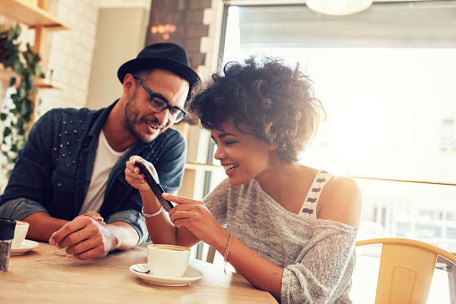Portrait of happy young man and woman sitting together at a restaurant and looking at a mobile phone. Young friends looking at smart phone while sitting in cafe
