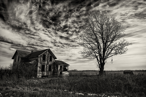 Black and white photo of an old scary abandoned farm house that is deteriorating with time and neglect.  The scene is enhanced with an old tree and a hangman's noose topped off with a dark, angry sky.