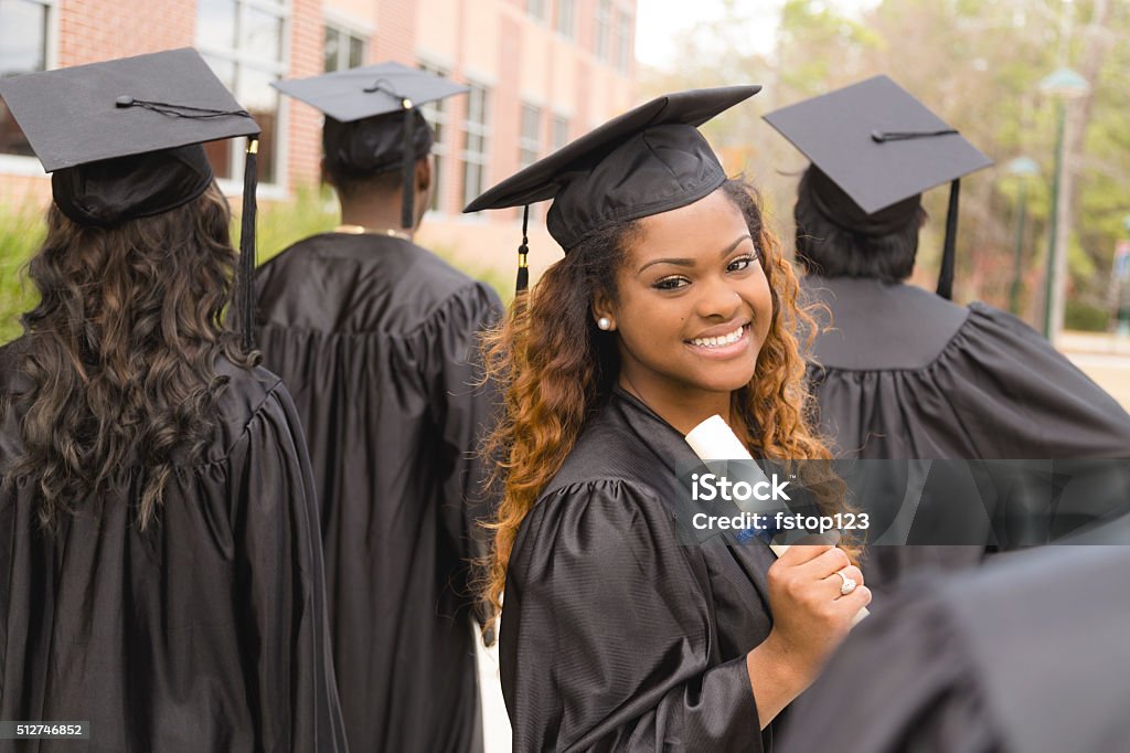 Education: African descent female graduate and friends on college campus. Candid moment of African descent female excitedly holding her diploma after the college graduation ceremony.  Her friends around her.  School building background on campus.  Graduation Stock Photo
