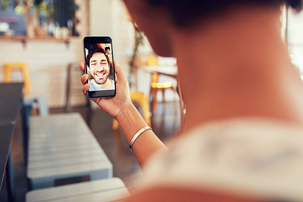 Woman having a video call with man on her phone Man and woman talking to each other through a videochat on a mobile phone. Woman having a video call with man on her smart phone. Woman sitting at a coffee shop. over the shoulder view photos stock pictures, royalty-free photos & images