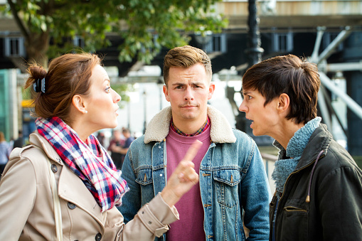 Young man dismayed as his girlfriend argues with ex outdoors. He looks at camera, frowning. Photographed in London, UK.