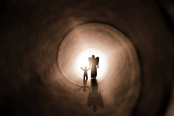 A concept image of a silhouette of an angel holding the hand of a small child as she walks him to a beautiful bright light at the end of a tunnel.