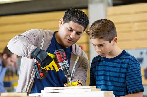 Mid adult Hispanic father or teacher in woodworking day camp show preteen son or student how to use a power drill in workshop. The boy watches as the man drills hole into the wood.  The man is wearing fingerless gloves.