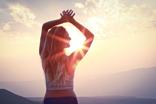 a long haired blonde female standing inferno of a sunset over the mountains with hands outstretched above head. sun is beaming through arms into a sun burst