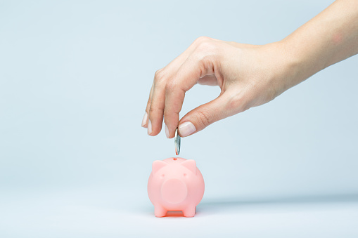 Female hand putting a coin into piggy bank. Light blue background, soft shadow. Canon 5D MK III