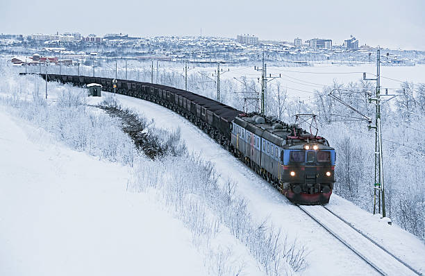 Electric locomotive hauling heavy iron ore train departing city (Kiruna) in Arctic Circle winter.  The wagons are loaded with tonnes of iron ore heading for a nearby seaport and export to Europe.  Horizontal, copy space, some grain due to low light and high ISO, ID removed.