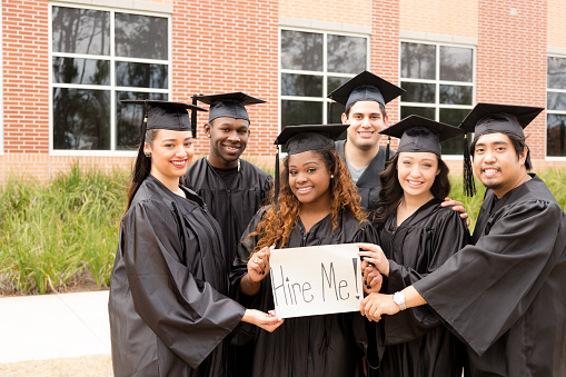 Six multi-ethnic graduates hold 'Hire Me' sign after college graduation. School building background. Employment issues concept.