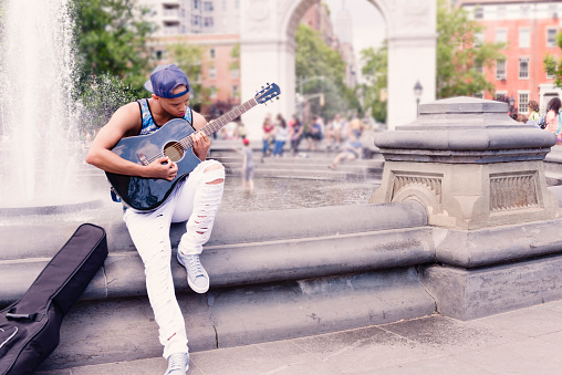 This is a color, royalty free, stock photograph of a smiling young Black man in his 20s playing the acoustic guitar outdoors on a summer day in NYC's Washington Square Park. Photographed  with a Nikon D800 DSLR camera.