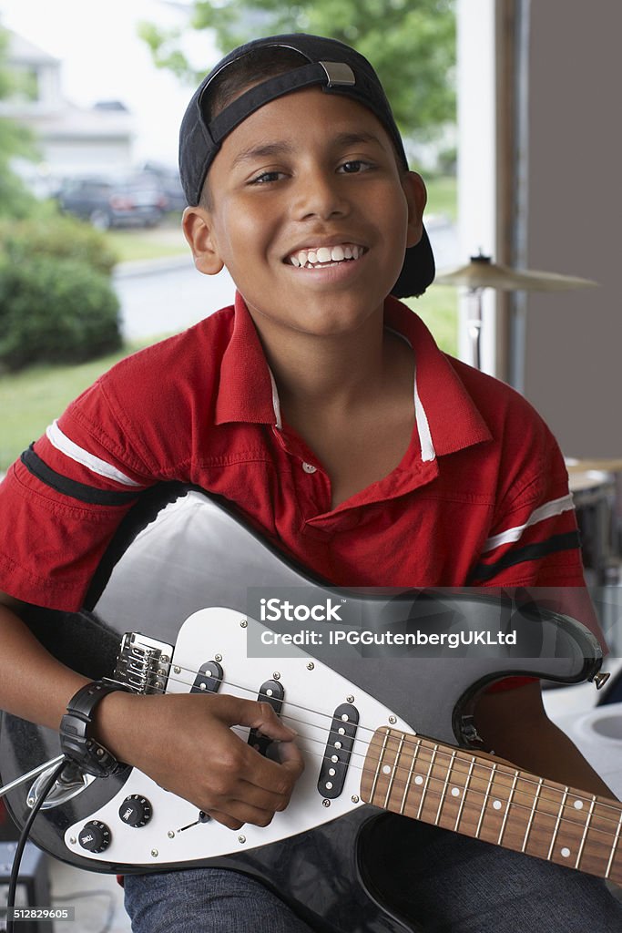 Boy Playing Guitar In Garage Portrait of smiling young boy playing electric guitar in garage Cap - Hat Stock Photo