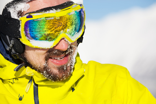 Close-up portrait of a man wearing beard ski mask and helmet with snow on face and mountain on background