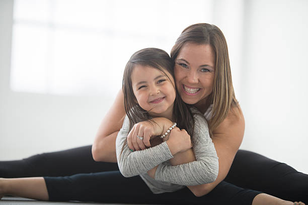 Mother and Daughter Stretching on the Mat A mother and daughter are doing yoga together on mother's day. They are both smiling and looking at the camera. long sleeved recreational pursuit horizontal looking at camera stock pictures, royalty-free photos & images