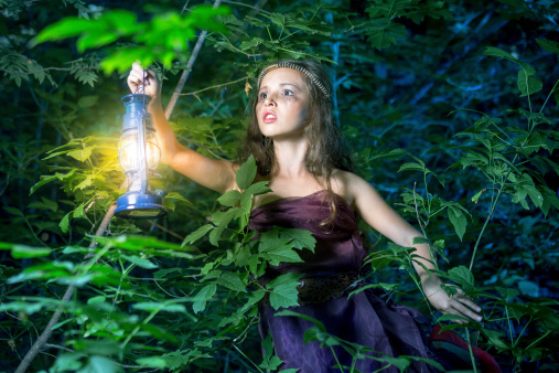 Charming teen girl princess standing with a lantern in hand at night forest