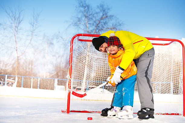 Father teach son to play ice hockey and hold hockey stick standing near gates on ice