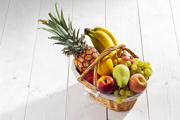 Fruit basket with pineapple, bananas, lemon, apple, peaches, grapes on white wooden background