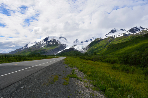A beautiful back road of Alaska leads up to green covered mountains with ice glaciers at their peaks seemingly cascading their beauty onto the roadway with nature's delight.
