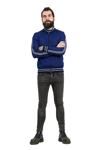 Confident proud bearded man in blue tracksuit jacket and tight jeans looking at camera with crossed arms. Full body length portrait isolated over white studio background.