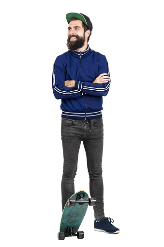 Young hipster in blue tracksuit jacket and baseball cap on skateboard looking away smiling. Full body length portrait isolated over white studio background.