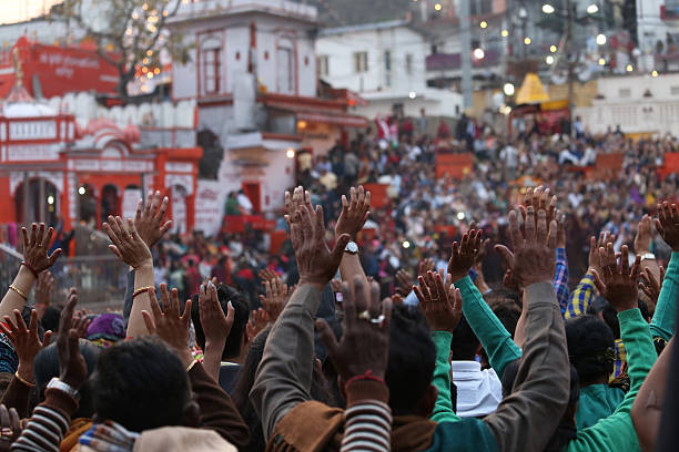 Rear view of Pilgrims praying during evening aarti at Har Ki Pauri, Uttarakhand, Haridwar. The Ganga Aarti (prayer) that takes place at Har Ki Pauri every evening after sunset is popular amongst pilgrims and tourists all over the world. The priests perform the Ganga Aarti at the ghat amidst thousands of devotees. People gather on both sides of the banks of the river to sing its praises. The priest stand on the ghat holding large fire bowls and the gongs in the temples start ringing. The air is filled with the fragrance of incense, sounds of various gongs/bells and the chants sung together by the priests and a large number of people present there.