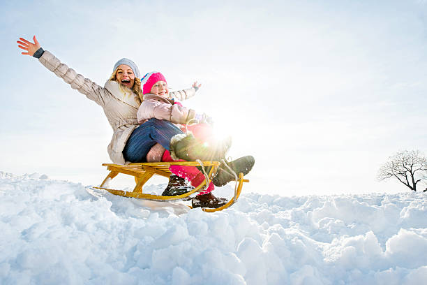 Mid adult mother and her daughter sledging in the snow and having fun. They are looking at the camera.  