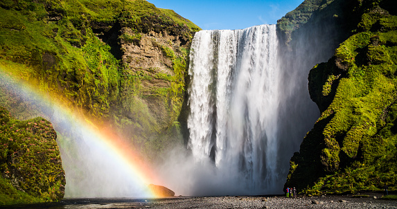 Skogafoss waterfall with beautiful rainbow. Rocks with lush green foliage. Tiny visitors in the foreground - Iceland