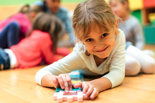 Portrait of Happy Little Girl in kindergarten Group of children in kindergarten. Showing their imagination and skill and creativity. Focus to little girl lying down, looking at camera and playing with colorful blocks. Kids around are playing. long sleeved recreational pursuit horizontal looking at camera stock pictures, royalty-free photos & images