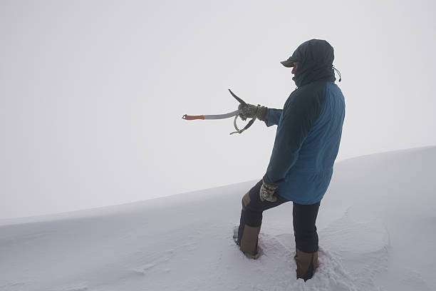A man is pointing with an ice axe at the top of a snowy mountain he just climbed