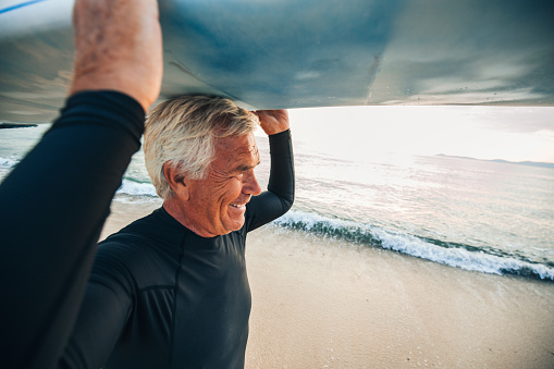 Portrait of a smiling senior surfer coming out of the water after a good surfing session, holding his surfboard over his head 
