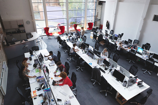 A horizontal image of a large office space shot from above. 10 office workers can be seen below. There are three large tables spread out across the room with multiple workstations.