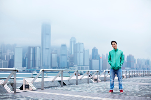 Full length portrait of a handsome young asian man standing in ftont of the Hong Kong skyline