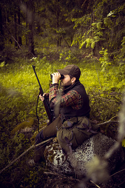 A mid aged man is sitting on a stone in a forest and observing the environment in the distance using binoculars. He also has a shotgun. He is wearing a cap, checkered shirt and warm waistcoat. His backpack is laying besides him. Image is dark and contrasty. Copy space available. Made in Northern Europe.