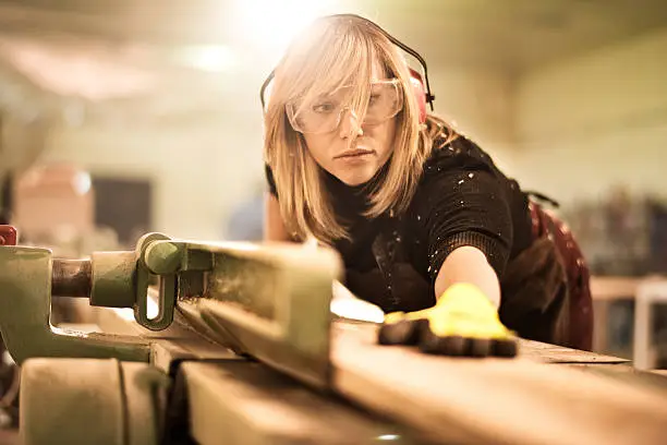 Blonde woman with protective equipment working with planks in workshop. She is making furniture parts.