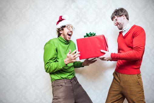 Two goofy looking men in ugly Christmas colored turtlenecks and sweaters exchange a holiday gift  Damask style vintage wall paper in the background.  Horizontal with copy space.
