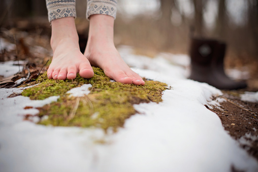 Child's bare feet, standing on a patch of moss, surrounded by snow, with boots in the background.