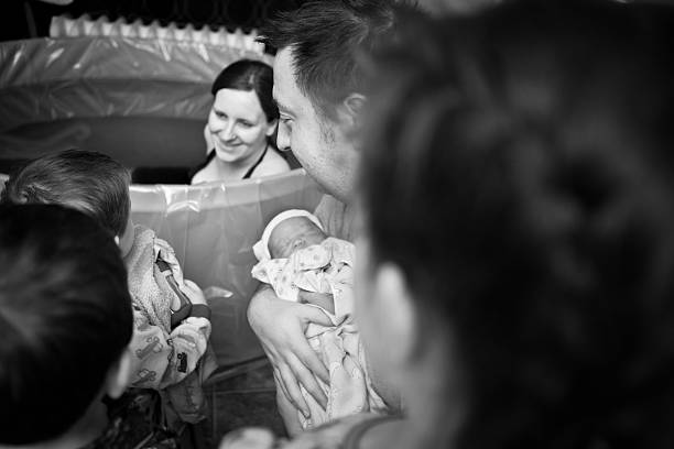Siblings Meeting Newborn Brother After Home Water Birth Black and white image of a father showing his newborn son to his other children as their happy mother looks on. water birth stock pictures, royalty-free photos & images