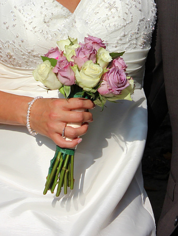 Photo showing a newy married bride wearing a traditional white wedding dress with a sweetheart neckline and diamante gems providing sparkling detail.  The bride is pictured holding her simple bridal bouquet, made from pale pink / lilac roses and cream rose flowers.  A pearl bracelet can be seen on the bride's wrist.