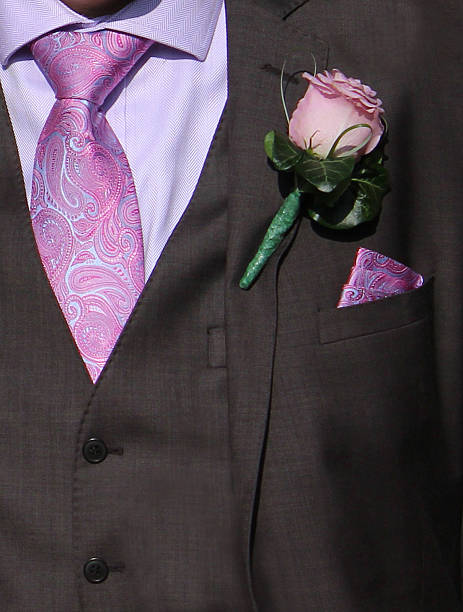Photo showing the detail of a bridegroom's smart outfit at his wedding.  He is pictured wearing a dark grey suit with a purple / pink rose buttonhole, a lilac shirt and a matching pink tie and handkerchief in his top pocket.  The bridegroom is just about to walk into the church, to wait for his bride to arrive and then get married.