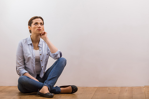 Attractive dreamy young woman in casual clothes looking away while sitting on wooden floor against white background