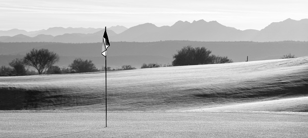 A beautiful desert golf course backlit. Phoenix, Arizona. A beautiful black and white image of a golf course in the sonoran desert in Arizona. Flag is backlit with beautiful rolling green and contours. Nobody is in the panoramic image, taken near Scottsdale. Themes include golf, resorts, summer, sports, leisure, mountain golf, desert golf, and the sonoran desert.
