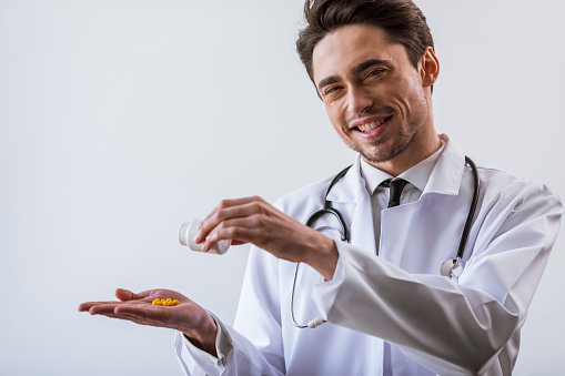 Handsome young doctor in white gown with stethoscope smiling, holding a bottle of medicine and pouring pills on his palm, on white background