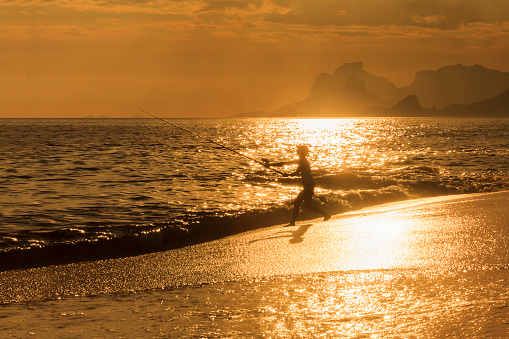 A DSLR photo of a woman using her fishing rod to trow a bait out in the sea in Camboinhas beach in Niteroi city, Rio de Janeiro, Brazil. She is silhouetted against a bright sun reflection over the sea and the wet sand. In the background are the mountains of Rio de Janeiro skyline.