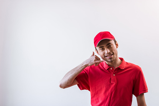 Handsome young worker in red t-shirt and cap smiling, looking at camera and imitating hand phone, on white background