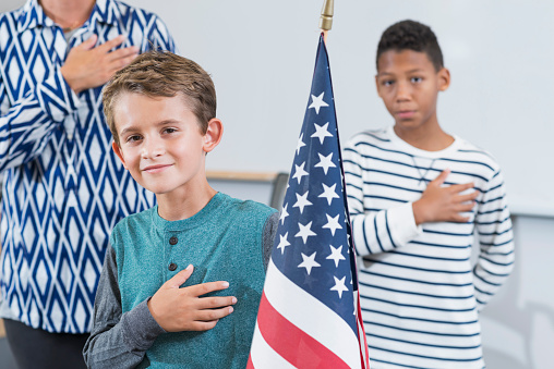 An elementary school student in the classroom saying the pledge of allegiance. The 9 year old boy is standing in front of the class holding the American flag, hand on his heart, smiling and looking at the camera. Another student and teacher are out of focus in the background.