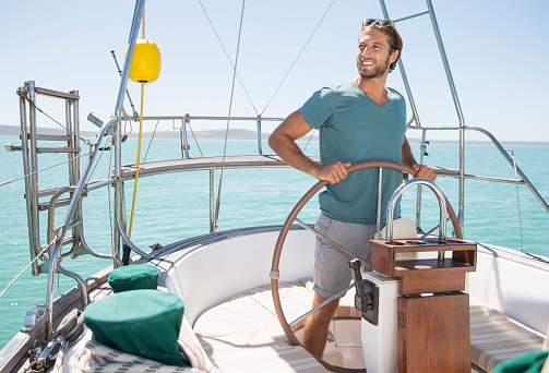 Waist up side shot with defocused background of a good looking sailor slightly smiling and looking at the horizon while holding firmly the boat helm enjoying his job on summer day in the Adriatic sea.