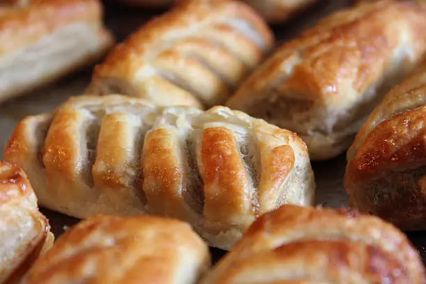 Photo showing a stainless steel oven tray of freshly baked sausage rolls cooking in the oven, with their puff pastry appearing golden brown after being glazed with an egg wash.