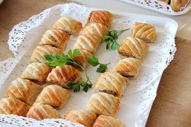 Photo showing a large plate of freshly baked, homemade sausage rolls, party food, just taken out of the oven with their puff pastry appearing golden brown after being glazed with an egg wash.  The sausage rolls are displayed on white paper doilies as part of a selection of help-yourself buffet food at a party.  A garnish of flat leaf parsley / coriander leaves has been added to the plate.