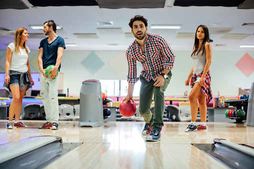 Male throwing bowling ball on bowling alley. Friends behind him looking at him. Four of them enjoy free weekend after hard working week. Location released.