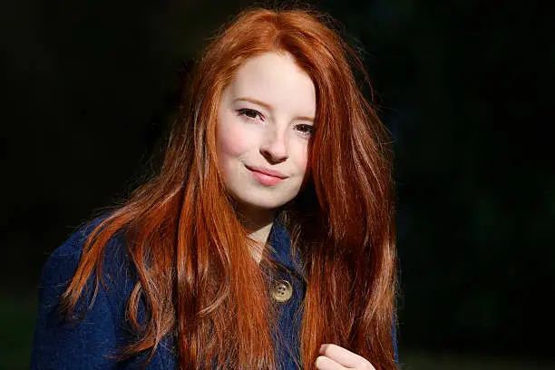 Photo showing a portrait of an attractive, teenage girl with long, flowing, red hair. This picture shows the teenager wearing a blue, winter coat against a blurred rhododendron backdrop.