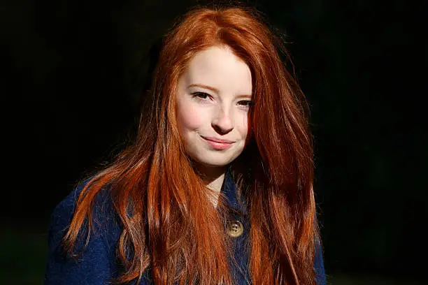 Photo showing a portrait of an attractive, teenage girl with long, flowing, red hair. This picture shows the teenager wearing a blue, winter coat against a blurred rhododendron backdrop.