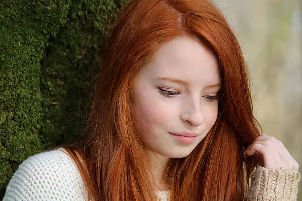 Photo showing a portrait of an attractive, teenage girl with long, flowing, red hair. This picture shows the teenager leaning against the trunk of a tree in a deciduous wood.